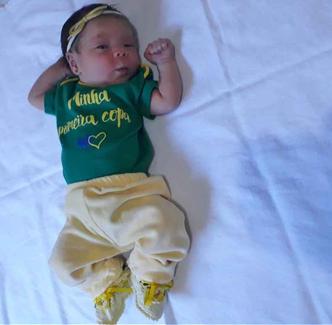 A pequena tem camiseta da Seleção Brasileira, e um body escrito A pequena tem camiseta da Seleção Brasileira, e um body escrito